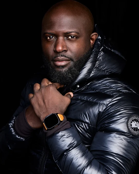 Portrait of Mario Rigby in a black Canada Goose down jacket, hand at his collar, wearing a watch with an orange strap.