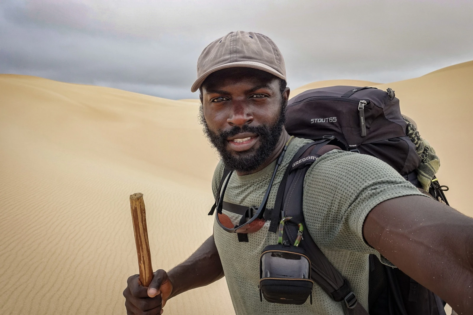 Mario Rigby carrying a backpack and walking stick among sand dunes, the photograph featured on his current deck cover.