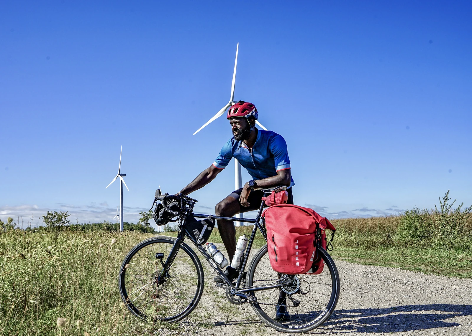 Mario Rigby with a loaded touring bicycle beside wind turbines.