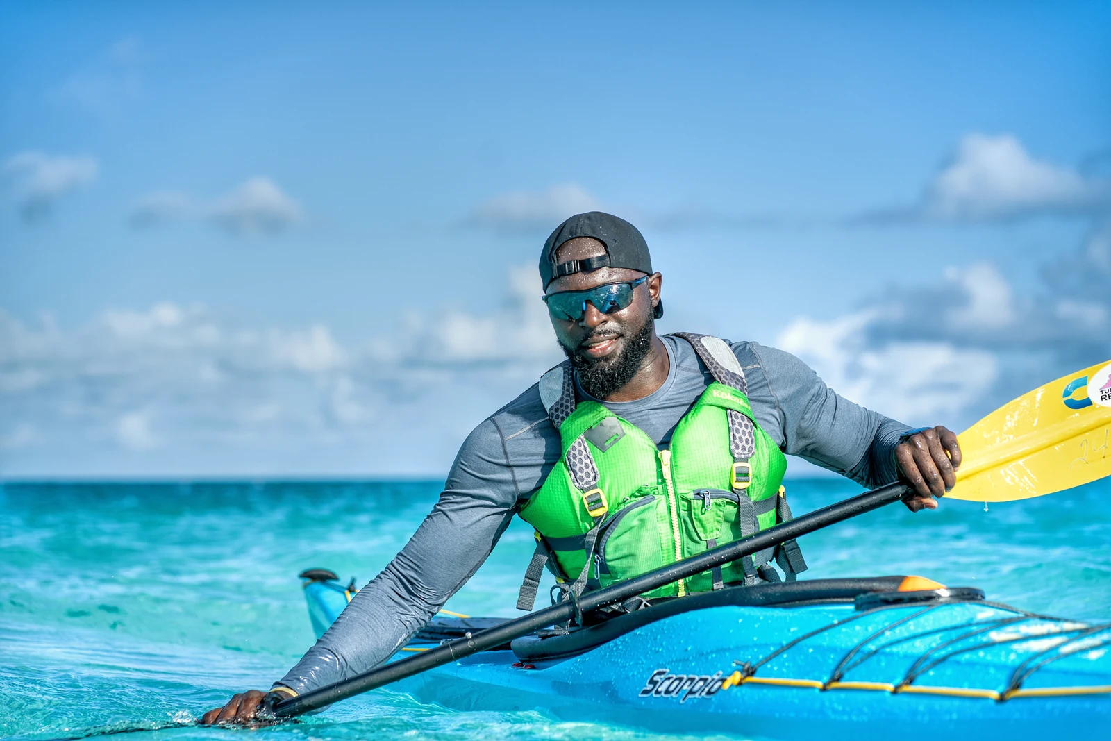 Mario Rigby seated in a kayak on turquoise water, holding a yellow paddle.