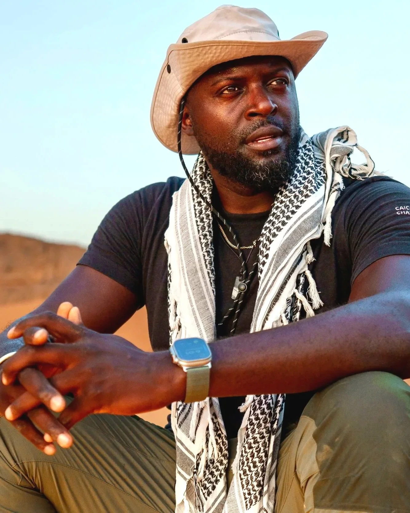 Mario Rigby seated in the desert, wearing a hat and patterned scarf.